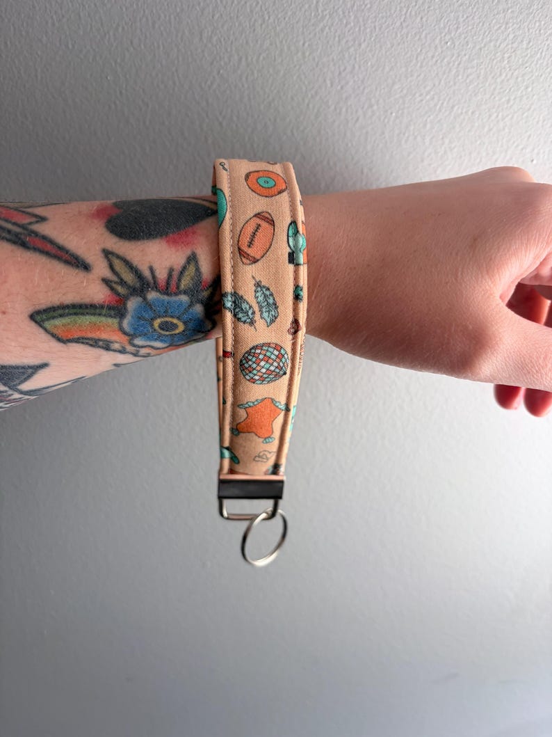 May include: A tan wristlet key fob with a repeating pattern of footballs, feathers, and other small illustrations. The key fob has a black metal clasp and a silver key ring.