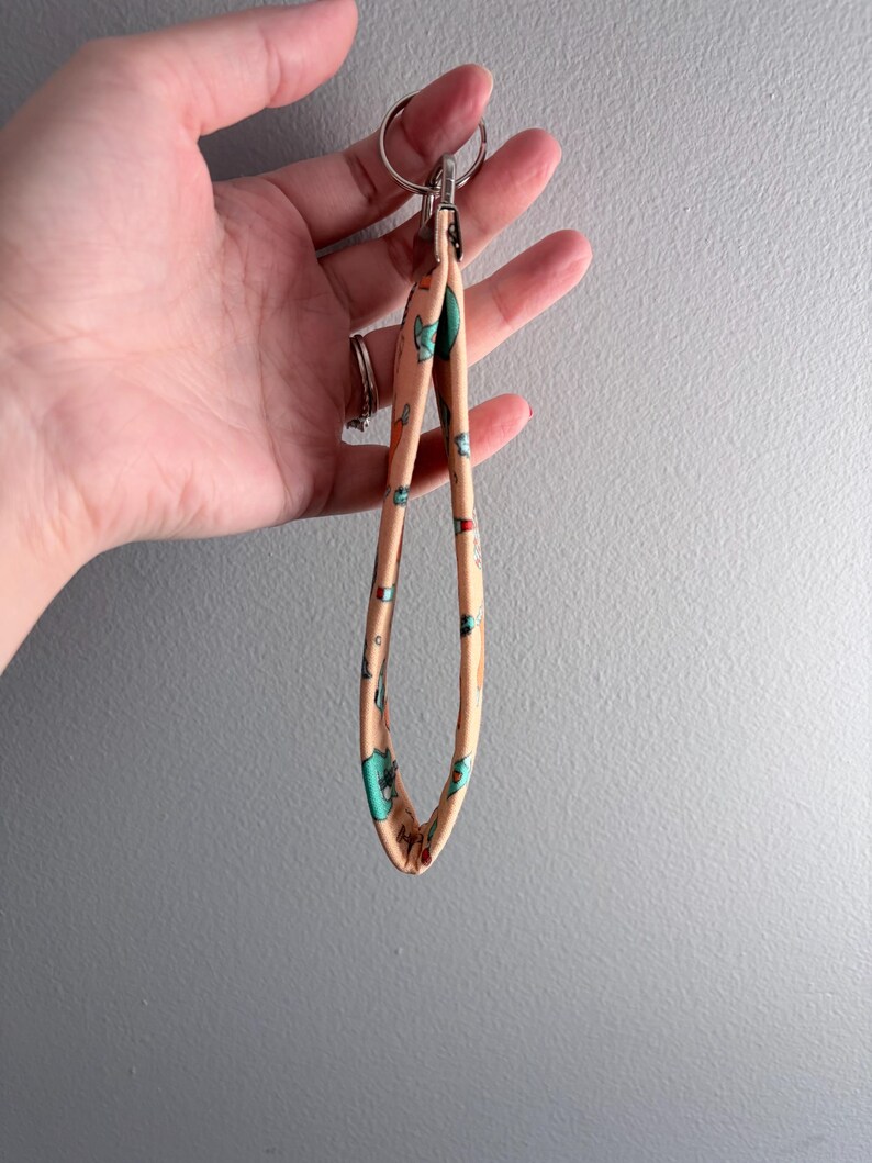 May include: A peach-colored wristlet keychain with a repeating pattern of cartoon animals in teal, blue, and red. The keychain has a silver metal clasp and a split ring for attaching keys. The wristlet is held in a hand.
