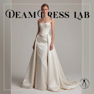 May include: A strapless ivory wedding gown with a fitted bodice and a flowing skirt. The bodice features vertical pleats, and the skirt has a dramatic train. The text "DREAM DRESS LAB" is displayed at the top.