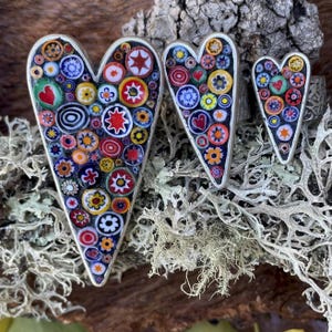 Peut inclure: Trois pendentifs en forme de cœur avec un motif de mosaïque. Les pendentifs sont remplis de morceaux de verre colorés de différentes formes et tailles, dont des cercles, des étoiles et des cœurs. Les pendentifs ont une bordure argentée et sont présentés sur du bois.