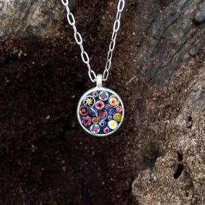 Puede incluir: Un collar de plata con un colgante redondo. El colgante presenta un diseño de mosaico colorido con patrones florales y de corazón rojos, amarillos, azules y blancos. La cadena es una delicada cadena de plata.