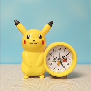 May include: A yellow Pikachu-shaped alarm clock with a round clock face. The clock face features black numbers, black hands, and small character illustrations. The clock is on a light-coloured surface against a blue background.