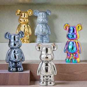 May include: A collection of six bear-shaped figurines in various metallic finishes. The figurines include gold, silver, gray, and iridescent colors. They are displayed on a wooden shelf, showcasing their glossy surfaces and unique designs.