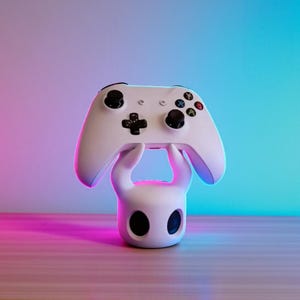 May include: A white gaming controller rests on a matching white stand. The controller has black buttons and colored action buttons. The stand has a unique design with two large, black eye-shaped cutouts. The background has a gradient of pink, purple, and blue.