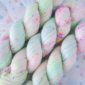 May include: A skein of soft yarn featuring a white base with pastel mint green and light pink sections. The yarn is speckled with vibrant dots of hot pink, bright blue, and yellow, and contains subtle metallic fibers that add a light shimmer.