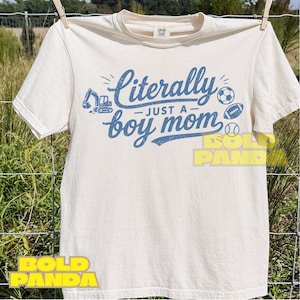 Puede incluir: Camiseta color crema con el texto azul "Literally just a boy mom". El diseño incluye ilustraciones de una excavadora, un balón de fútbol, un balón de rugby y una pelota de béisbol. La camiseta está colgada en un tendedero.