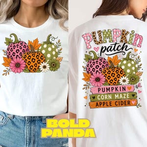 May include: A white t-shirt featuring a graphic of three pumpkins with leopard and polka dot patterns, surrounded by flowers and leaves. The back design includes the text PUMPKIN patch, PUMPKIN, CORN MAZE, and APPLE CIDER on colourful wooden signs.