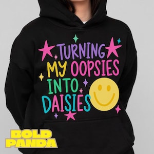 May include: A black hooded sweatshirt featuring a colorful graphic print that reads "TURNING MY OOPSIES INTO DAISIES" in purple, yellow, green, and pink lettering, accented by pink stars and a large yellow smiley face icon on the front.