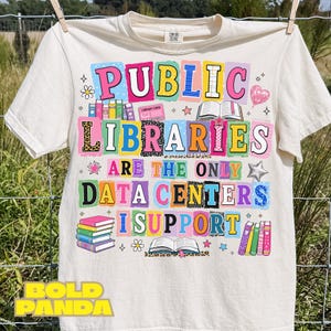 Könnte beinhalten: Ein cremefarbenes Kurzarm-T-Shirt mit einem bunten Grafikdruck und dem Text "PUBLIC LIBRARIES ARE THE ONLY DATA CENTERS I SUPPORT". Das Design enthält Illustrationen von gestapelten Büchern, einem aufgeschlagenen Buch und einem Bibliotheksausweis.