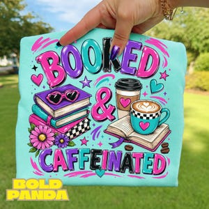 Puede incluir: Una camiseta azul claro con un estampado gráfico que incluye el texto "BOOKED & CAFFEINATED". El diseño muestra una pila de libros con gafas en forma de corazón, un vaso de café, un latte, flores rosas, estrellas y corazones en rosa y turquesa.