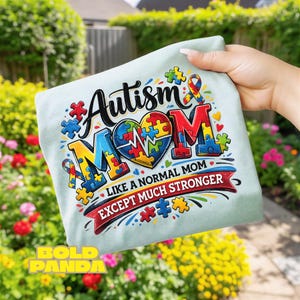 May include: A light blue t-shirt with the words "Autism Mom" in black and red, with a heart-shaped puzzle design. The text "Like a normal mom except much stronger" is below. The shirt is held up in front of a garden.