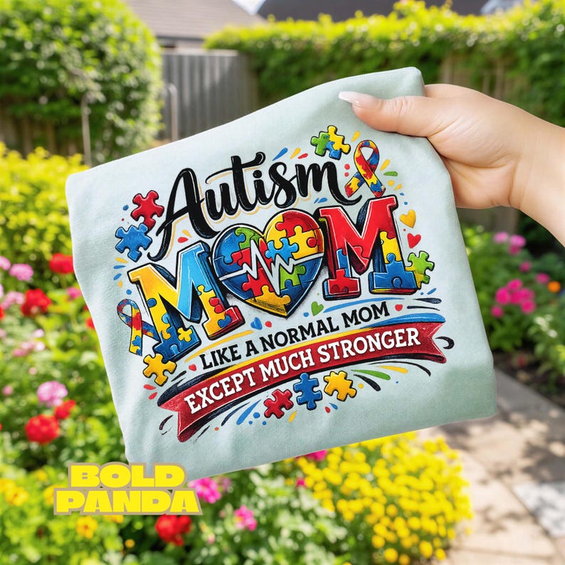 May include: A light blue t-shirt with the words "Autism Mom" in black and red, with a heart-shaped puzzle design. The text "Like a normal mom except much stronger" is below. The shirt is held up in front of a garden.