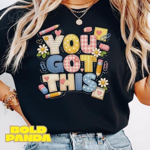 May include: Black t-shirt with the phrase "YOU GOT THIS" in colorful, patterned letters. The design includes pencils, erasers, hearts, and flowers, creating a cheerful, school-themed graphic.