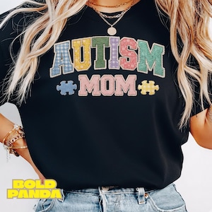 Puede incluir: Camiseta negra con las palabras "AUTISM MOM" en letras coloridas estilo patchwork y gráficos de piezas de rompecabezas. La camiseta es de cuello redondo y está hecha de un material suave.