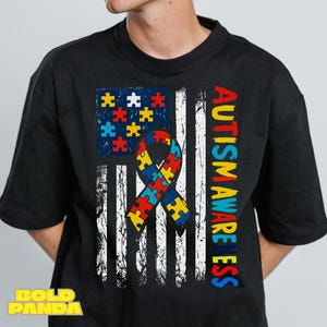 May include: Black t-shirt featuring a distressed American flag design with a puzzle piece pattern and a ribbon. The words "AUTISM AWARENESS" are printed vertically in colorful letters. The shirt has a crew neck.
