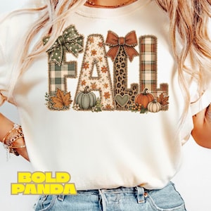 May include: A cream-coloured t-shirt featuring the word FALL in large block letters. The letters are decorated with green plaid, floral, leopard print, and brown plaid patterns, accented with bows, pumpkins, and autumn leaves.