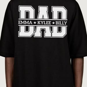 May include: Black t-shirt featuring the word "DAD" in large white block letters. Underneath, the names "EMMA", "KYLEE", and "BILLY" are displayed in white. The shirt has short sleeves.