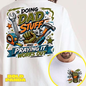 May include: A white t-shirt featuring a graphic print of tools including a wrench, hammer, screwdriver, and tape measure with the text "DOING DAD STUFF AND PRAYING IT WORKS OUT". A smaller version of the graphic with a gloved hand and wrench is on the front.