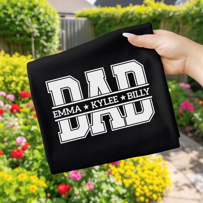 May include: Black fabric featuring the word "DAD" in large white block letters. Underneath, the names "EMMA * KYLEE * BILLY" are displayed in white. The fabric is held up by a hand.