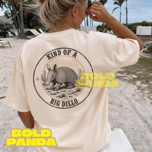 May include: Cream-colored t-shirt featuring a circular graphic with an armadillo, cactus, and the text "KIND OF A BIG DILLO." The design is in black and tan tones. The shirt is a casual, unisex style.