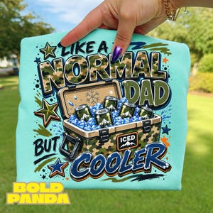 May include: A light blue t-shirt featuring a graphic of a camouflage cooler filled with ice and beverage cans. The text reads: LIKE A NORMAL DAD BUT COOLER, with ICED printed on the cooler. The design includes star accents and paint splatters.