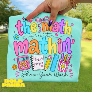 May include: A light blue t-shirt featuring the colorful text "the math Ain't mathin'" and "Show Your Work" with illustrations of a pink calculator, a notebook with math equations, a yellow pencil, a blue ruler, and a cup of pens.