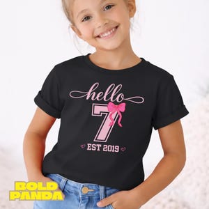 May include: Black t-shirt with the text "hello 7 EST 2019" in pink. The number 7 is in a pink box with a pink bow. The shirt is worn by a young person.