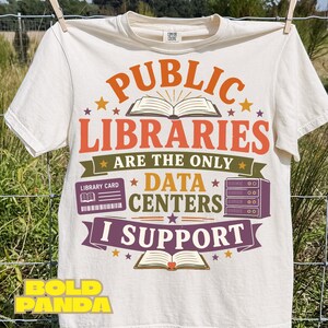 May include: An off-white short-sleeve t-shirt featuring a graphic with the text PUBLIC LIBRARIES ARE THE ONLY DATA CENTERS I SUPPORT. The design includes illustrations of an open book, a library card, and a server rack in orange, green, and purple.
