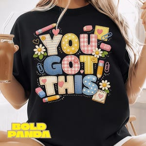 May include: Black sweatshirt with the phrase "YOU GOT THIS" in colorful, stitched-style letters. The design includes pencils, erasers, hearts, daisies, and paperclips, creating a cheerful, school-themed graphic.