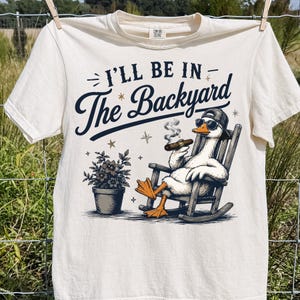以下が含まれることがあります： オフホワイトの半袖Tシャツ。野球帽とサングラスを着用し、ロッキングチェアに座って葉巻を吸うアヒルのグラフィックが描かれています。テキストは「I'LL BE IN The Backyard」とあり、鉢植えの植物が添えられています。