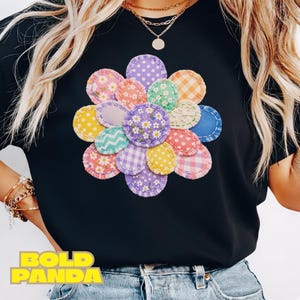Op de afbeelding: Zwart T-shirt met een bloemdesign gemaakt van stoffen patches in verschillende patronen en kleuren. De bloem bevat stippen, ruitjes en bloemenprints in roze, geel, paars en blauw. De tekst "BOLD PANDA" is zichtbaar aan de onderkant.