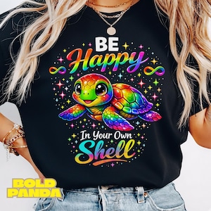 Puede incluir: Camiseta negra con un diseño colorido de una tortuga y el texto "BE Happy In Your Own Shell". La tortuga tiene una cara verde y ojos grandes. El diseño incluye estrellas y símbolos de infinito.