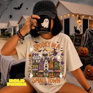 May include: A beige t-shirt featuring a colourful graphic of a spooky small town with a water tower, a witch on a broom, ghosts, and flowers. The text reads SPOOKY IN A SMALL TOWN and 689883. A black mug with a white ghost and the text BOO! is held.