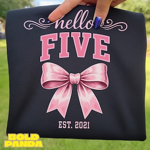 Puede incluir: Una camiseta negra con las palabras "hello FIVE" en rosa y un lazo rosa. El texto "EST. 2021" está debajo del lazo. La camiseta está sujeta por una mano.
