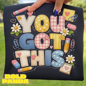 May include: A navy blue t-shirt with the phrase "YOU GOT THIS" in colorful, stitched-style letters. The design includes pencils, erasers, paperclips, hearts, and daisies. The words "BOLD PANDA" are at the bottom.