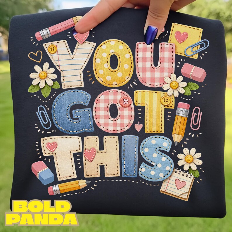 May include: A navy blue t-shirt with the phrase "YOU GOT THIS" in colorful, stitched-style letters. The design includes pencils, erasers, paperclips, hearts, and daisies. The words "BOLD PANDA" are at the bottom.