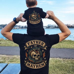 Puede incluir: Camisetas negras con diseños a juego. La camiseta de adulto presenta el texto "FATHER OF MAYHEM" con un gráfico de calavera y serpiente. La camiseta infantil tiene la palabra "MAYHEM" con un gráfico de calavera y serpiente. Ambas camisetas tienen un estilo de tatuaje vintage.