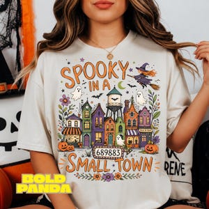 May include: A cream-coloured t-shirt featuring a colourful graphic of a small town with houses, a water tower, pumpkins, ghosts, and a witch on a broomstick. The text reads "SPOOKY IN A SMALL TOWN" and includes the number "689883" on a sign.