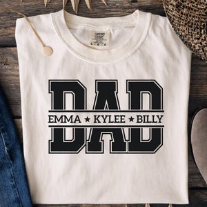 May include: A cream-coloured t-shirt featuring the word "DAD" in bold, black, block letters. Below the word "DAD" are the names "EMMA * KYLEE * BILLY" in black. The shirt is a Comfort Colors brand.