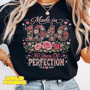 Op de afbeelding: Zwart T-shirt met de tekst "Made in 1946" in een bloemenontwerp. Het nummer 1946 is gevuld met roze bloemen en groene bladeren. Daaronder staat de tekst "80 Years Of Perfection".