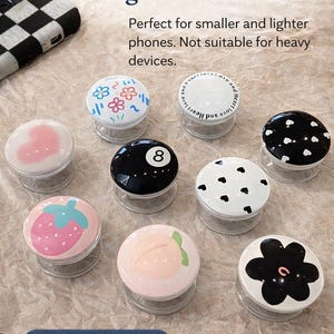 May include: Nine pop sockets with various designs, including flowers, hearts, a strawberry, and a peach. The text reads "Cute Pop Sockets for Lighter Phones" and "9 Cute Designs!" These are designed for smaller and lighter phones.