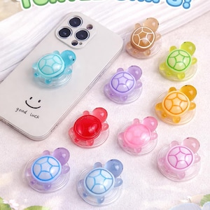 May include: A collection of colorful turtle-shaped phone grips. The grips are in various colors, including blue, green, purple, red, and pink. The text "CUTE TURTLE GRIPS!" is displayed at the top of the image.