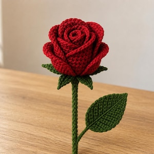May include: A handcrafted crochet rose featuring a deep red flower head with layered petals, a green calyx, a long green stem, and a single green leaf attached to the side. The texture of the yarn is visible throughout the entire structure.