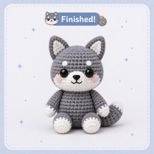 Peut inclure: Un chien Shiba Inu au crochet gris et blanc. La peluche a de grands yeux noirs, des joues roses et un museau blanc. Le texte "Finished!" est affiché au-dessus du chien.
