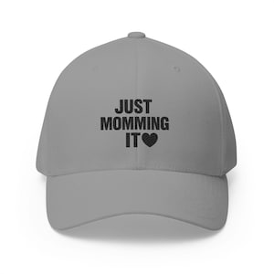 May include: A grey baseball cap featuring the embroidered black text "JUST MOMMING IT" followed by a black heart symbol centered on the front panel. The cap has a structured crown, a curved brim, and a button at the top of the dome.