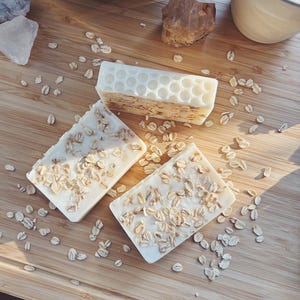 May include: Three bars of handmade soap with visible oatmeal flakes, arranged on a wooden surface. One bar has a honeycomb pattern. The soaps are off-white, and the oatmeal adds a textured appearance. Scattered oats surround the soaps.