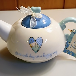 May include: A white ceramic teapot with a light blue lid, a heart-shaped knob, and a white ribbon. The body features a patchwork heart graphic with the word love and the text Start each day in a happy way. A Holly Hobbie tag is attached.