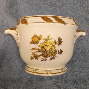 May include: A white ceramic bowl with gold floral and leaf designs. The bowl has two handles and a decorative gold trim around the top and base. The floral design features a large rose and smaller flowers.
