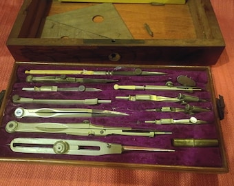 May include: A vintage wooden box with a purple velvet interior, housing a collection of antique drafting tools. The tools are made of metal and include various compasses, dividers, and other instruments. A wooden triangle and other pieces are in the lid.