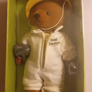 May include: A brown teddy bear wearing a yellow hard hat and a white jumpsuit with the text Shell Service printed on the chest. The bear is positioned inside a green cardboard box and holds a small grey tool in its right paw.